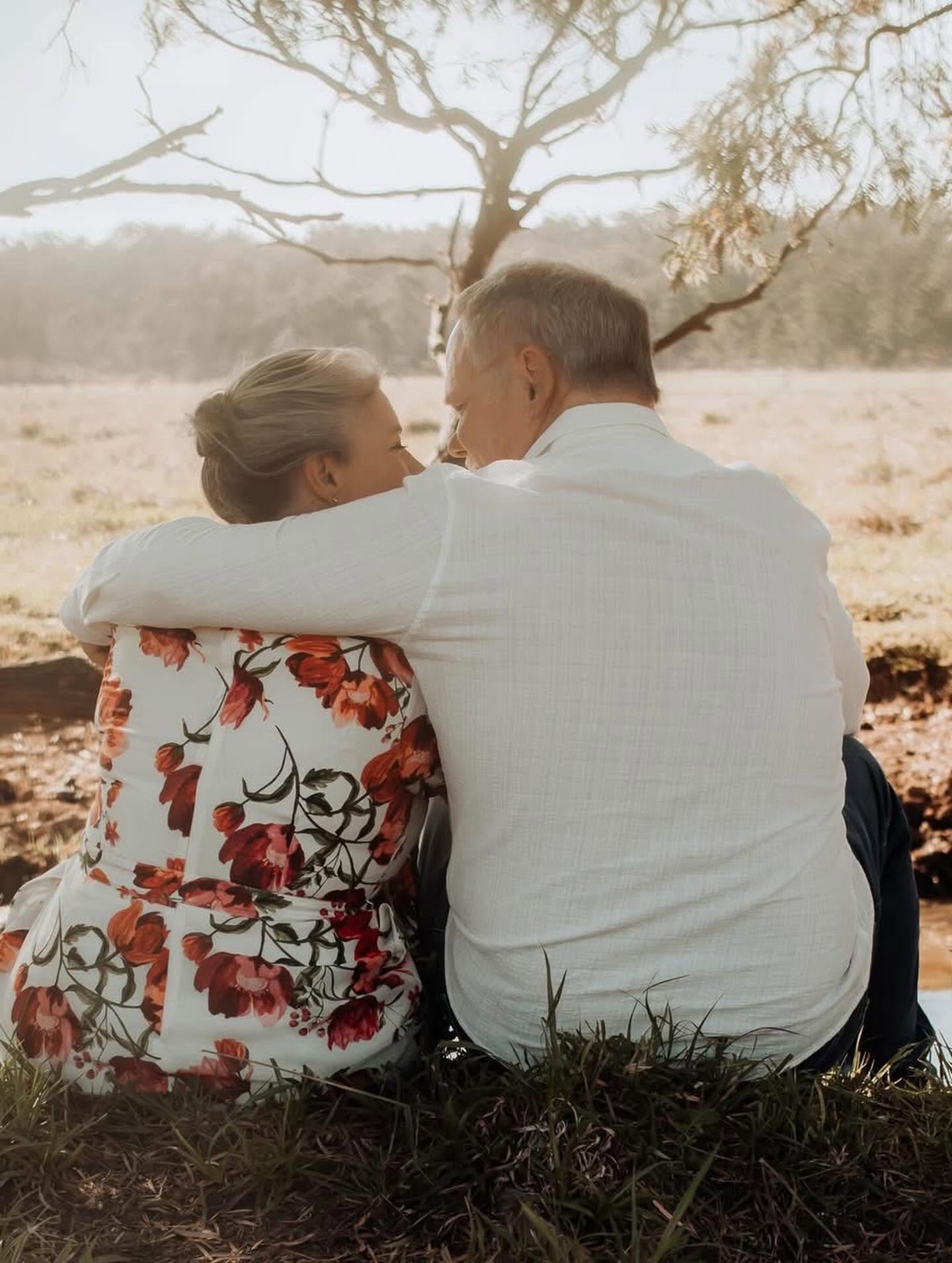 A couple sitting together beneath a gum tree, his arm around her shoulders